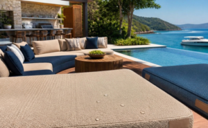 Sofá modular e espreguiçadeiras de luxo revestidos com material sintético náutico bege e azul em uma varanda gourmet com deck e piscina de borda infinita sob sol intenso. Ao fundo, uma lancha está ancorada no mar turquesa, integrando o paisagismo tropical e a arquitetura residencial de alto padrão.